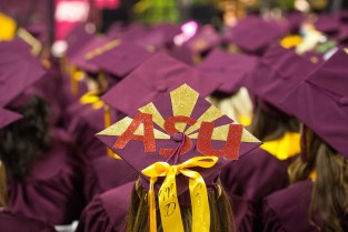 View from behind a group of graduating students in their caps and gowns. Featured is a mortarboard decorated with gold ribbons and the letters A S U.